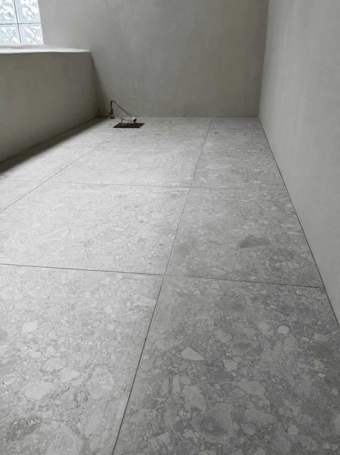 A large-format terrazzo-look stone tile floor meeting rounded microcement walls, lit by a stained-glass window.
