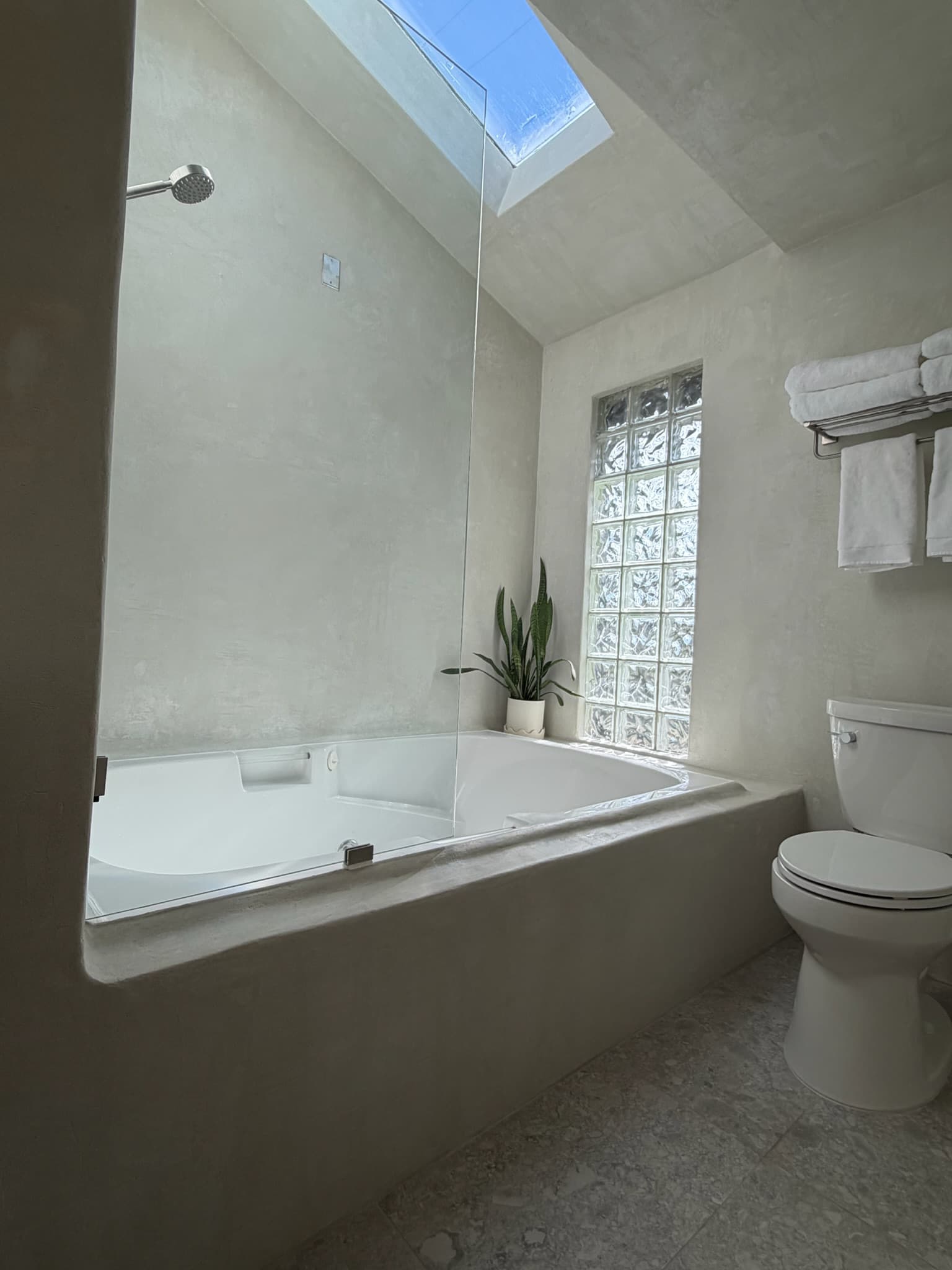A microcement bathroom with a soaking tub, a full-height glass shower panel, a skylight, and a glass-block window.