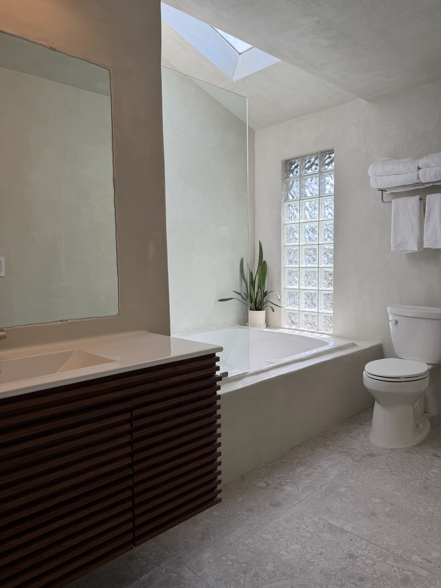 A floating walnut slat vanity beside a soaking tub in a warm microcement bathroom with a glass-block window.