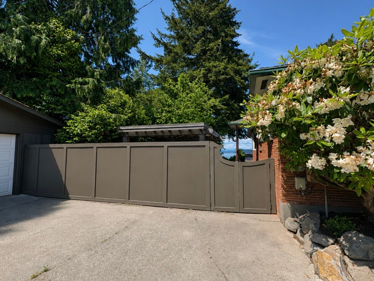 A tall gray board fence and custom gate with a small metal-roofed cover, framing a water view beside a brick home.